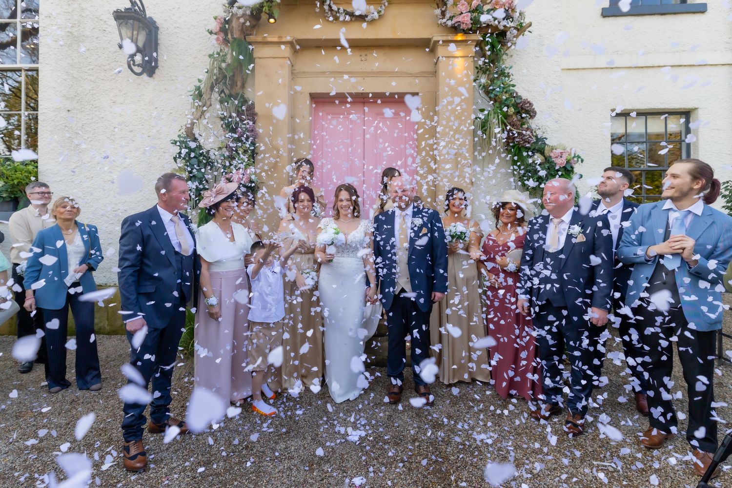 Wedding party celebrating with confetti outside a venue with a pink door and floral arch decoration. Casual Captures Wedding The Croft Hotel