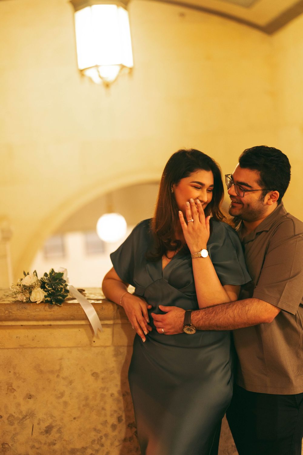 A couple shares a joyful engagement moment, woman covering her smile, man embracing her warmly in a warmly lit elegant venue.