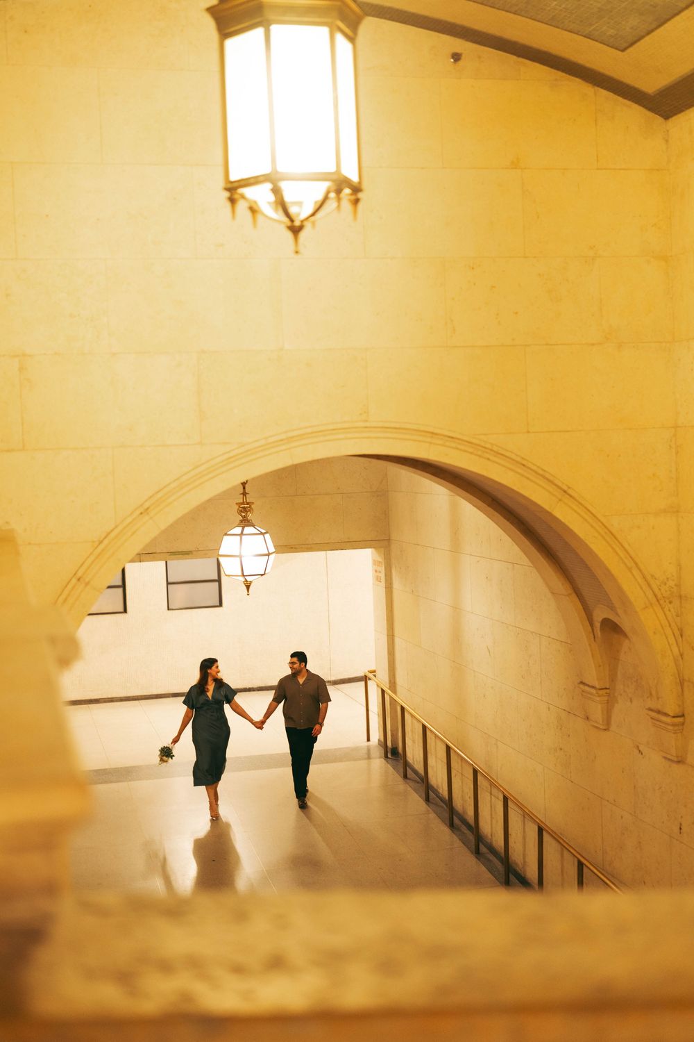 Couple holding hands walking through elegant arched hallway with warm golden lighting and vintage lanterns.