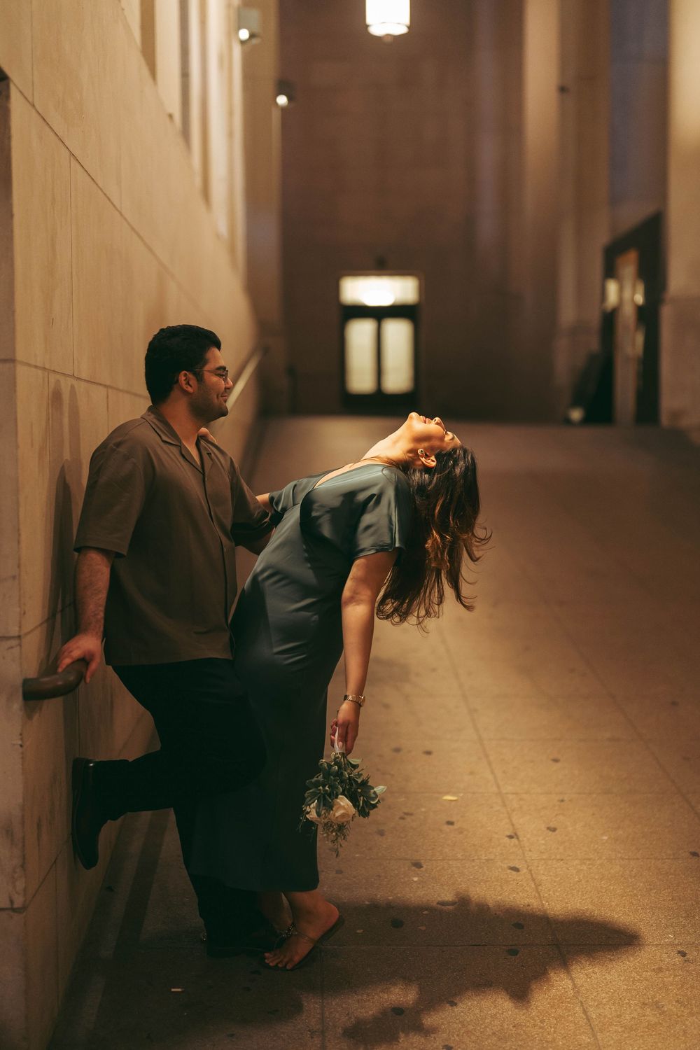 Couple posing in dimly lit hallway at night, woman leaning forward with bouquet, man against wall in urban setting.