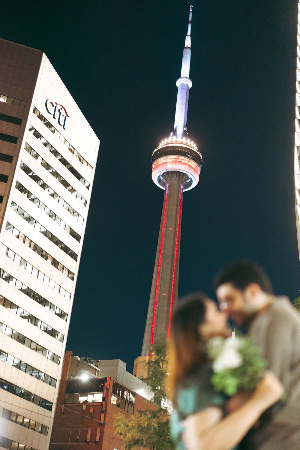 Couple kissing at night with the illuminated CN Tower and Citi building in Toronto skyline background.