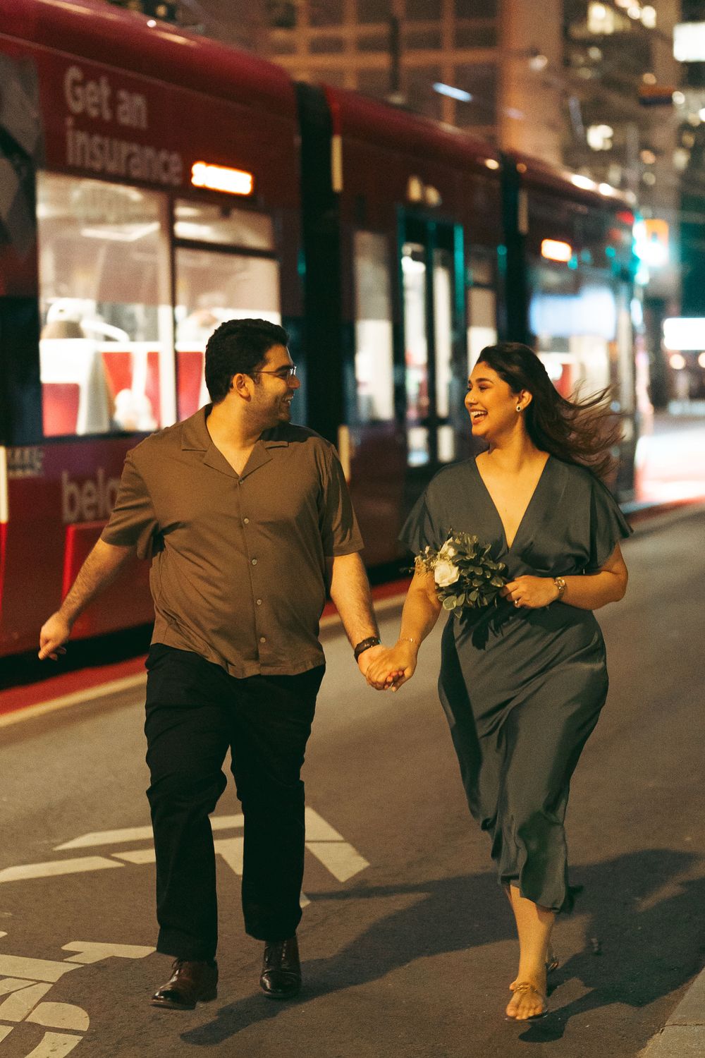 Couple holding hands walking at night on city street, woman in gray dress holding flowers, red bus in background.