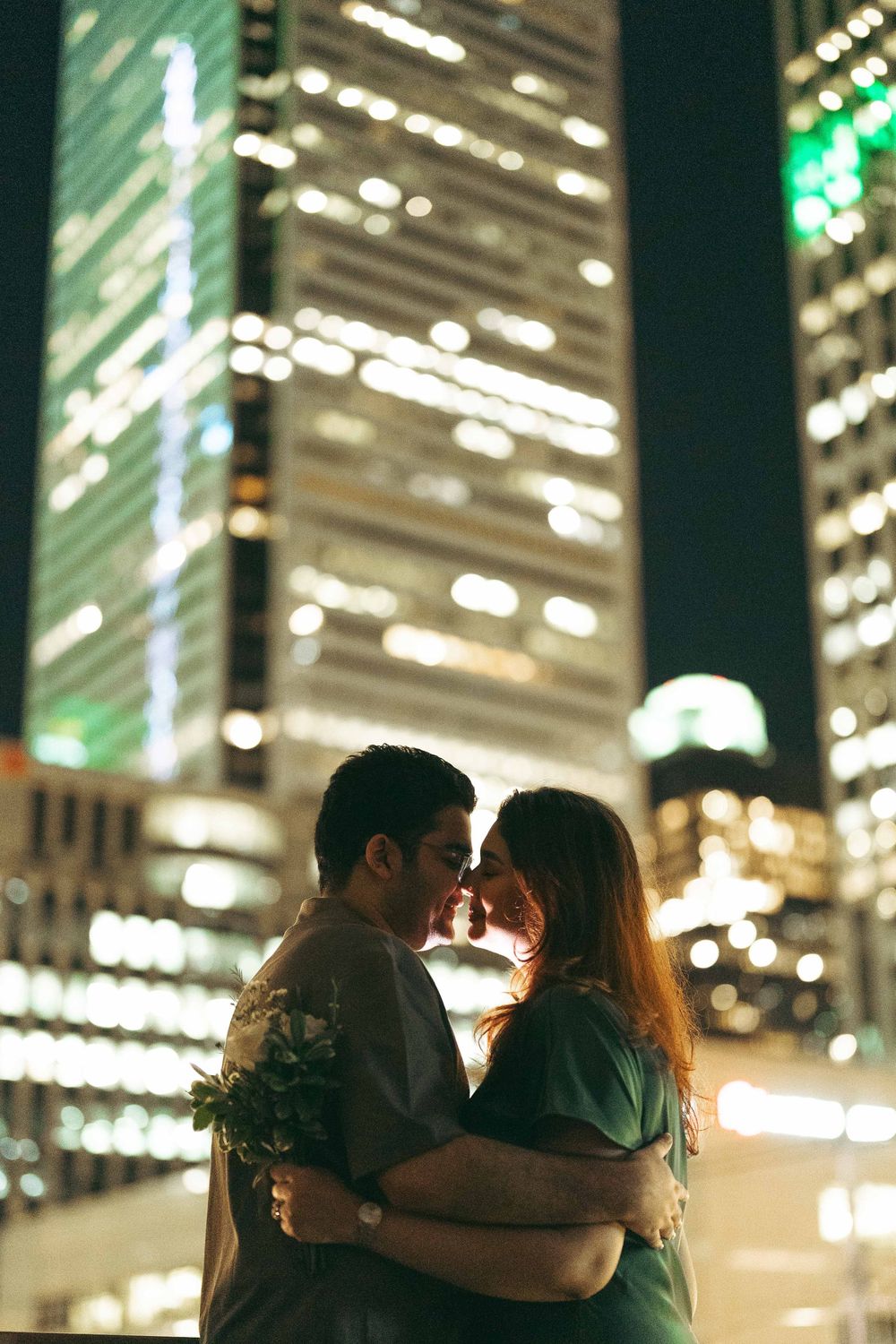 Romantic couple embracing and kissing in front of illuminated city skyscrapers at night.