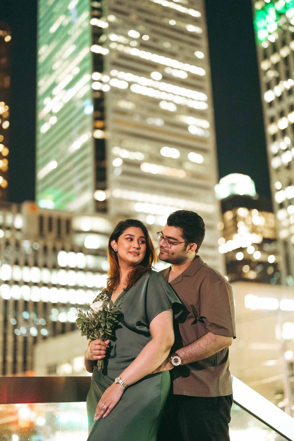 Couple posing romantically at night with illuminated city skyscrapers in background, woman holding green bouquet.