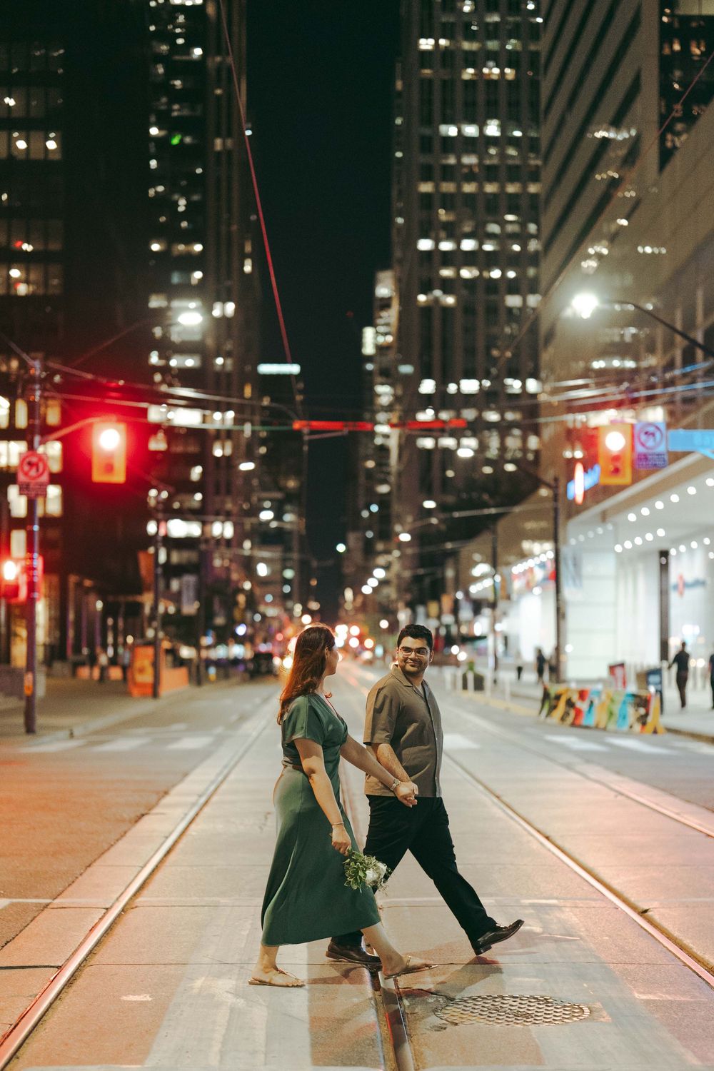 Couple holding hands crossing a city street at night with illuminated skyscrapers and traffic lights in the background.