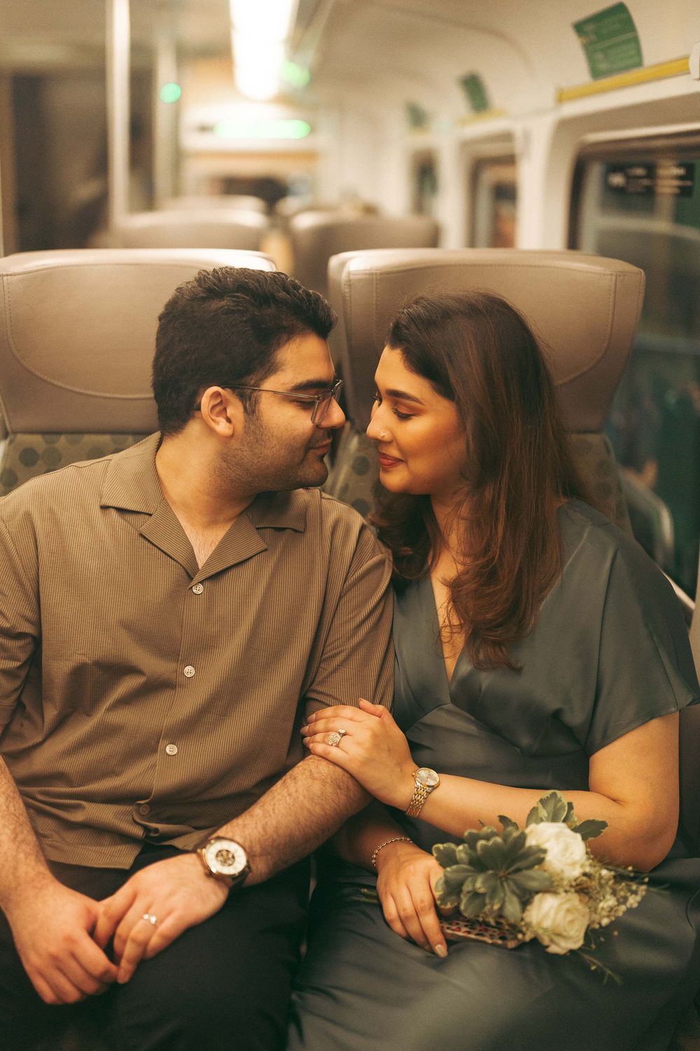 A couple sits together on a train, holding hands and gazing at each other, woman holding white flowers.