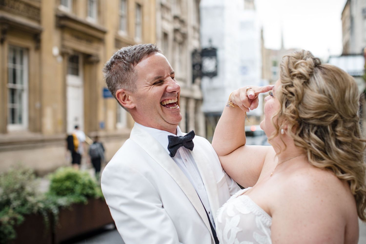 Joyful groom in white tuxedo laughing with bride in elegant updo on a city street wedding day.