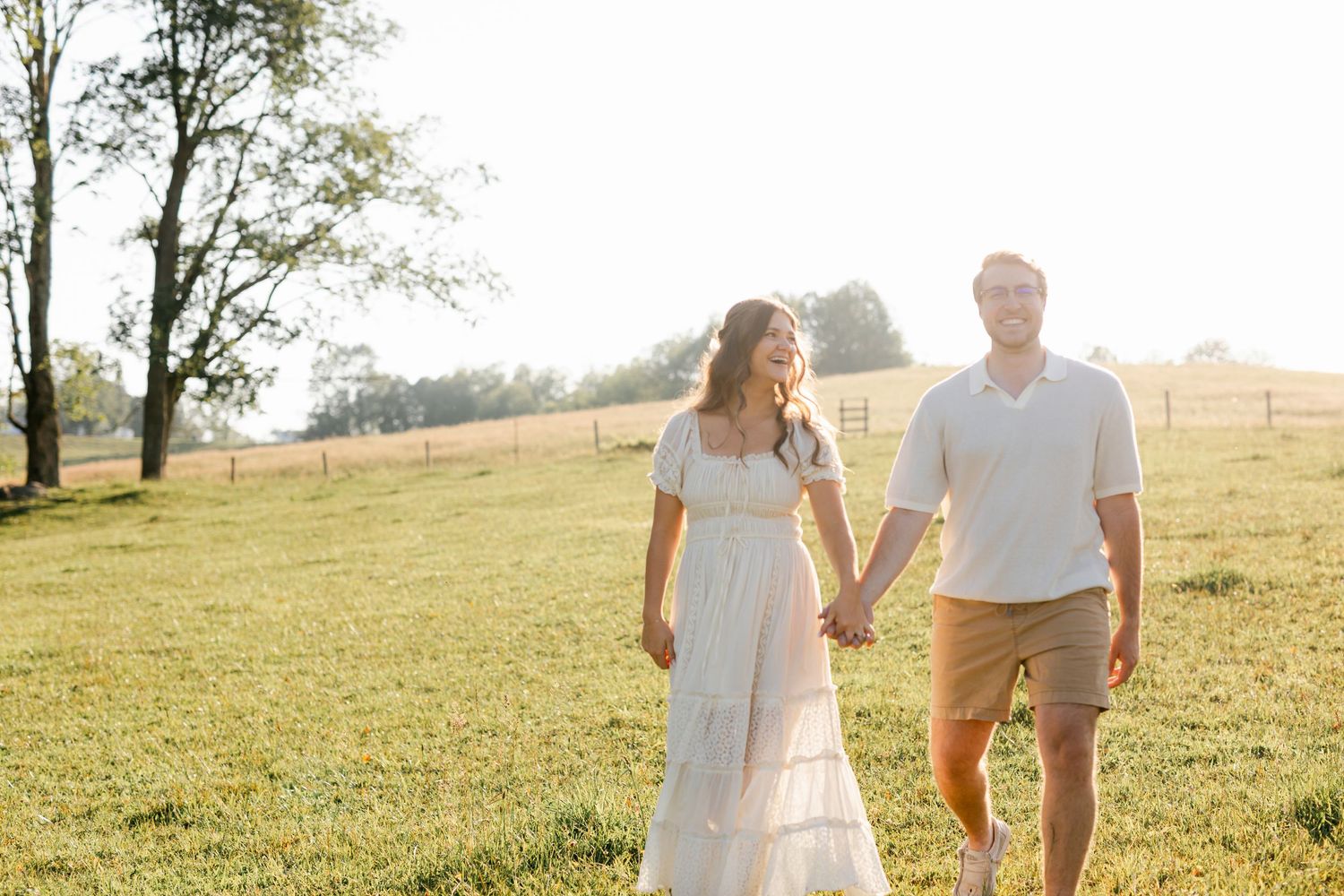 Couple walks hand in hand through golden sunlit field during outdoor engagement session.
