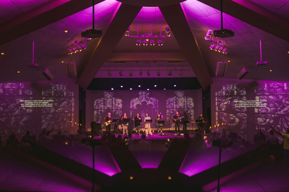 A church worship service bathed in purple lighting with a band performing on stage amid dramatic projected visuals.