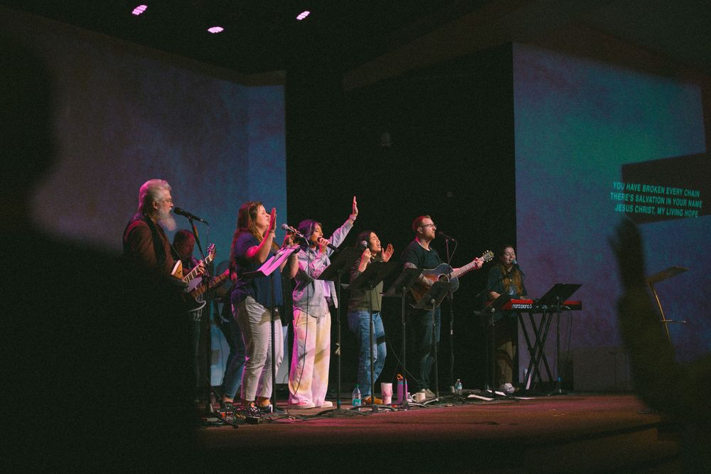 Worship team performs on colorful lit stage with lyrics displayed on screen behind them.