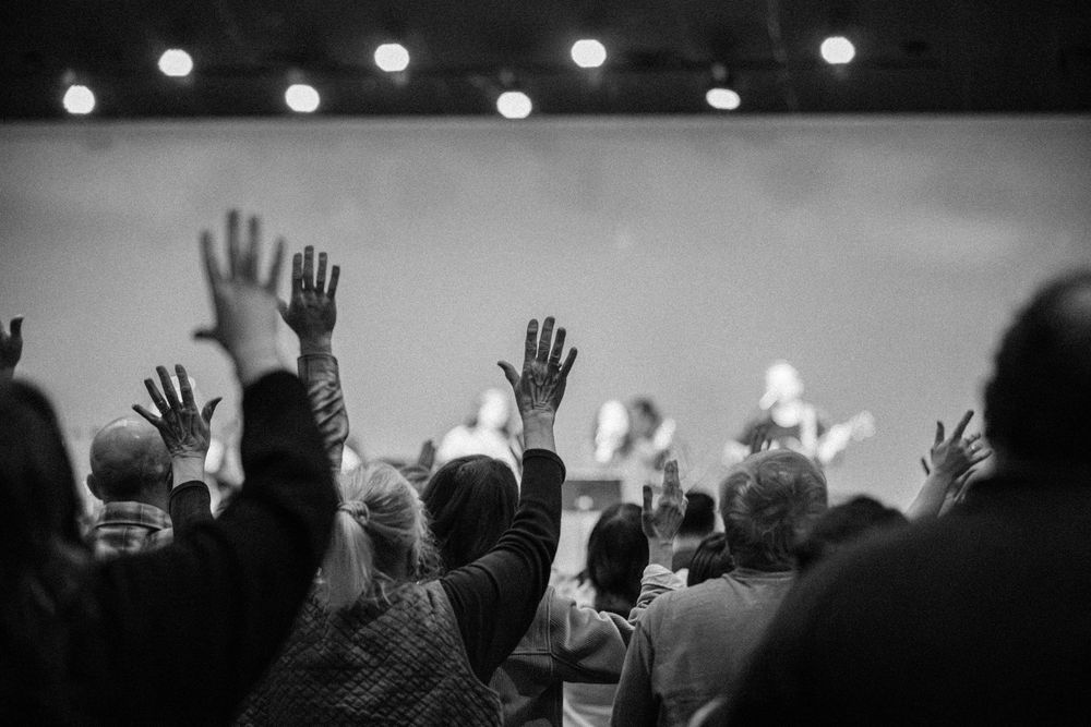 Black and white image of congregation raising hands toward performers on a lit church stage.