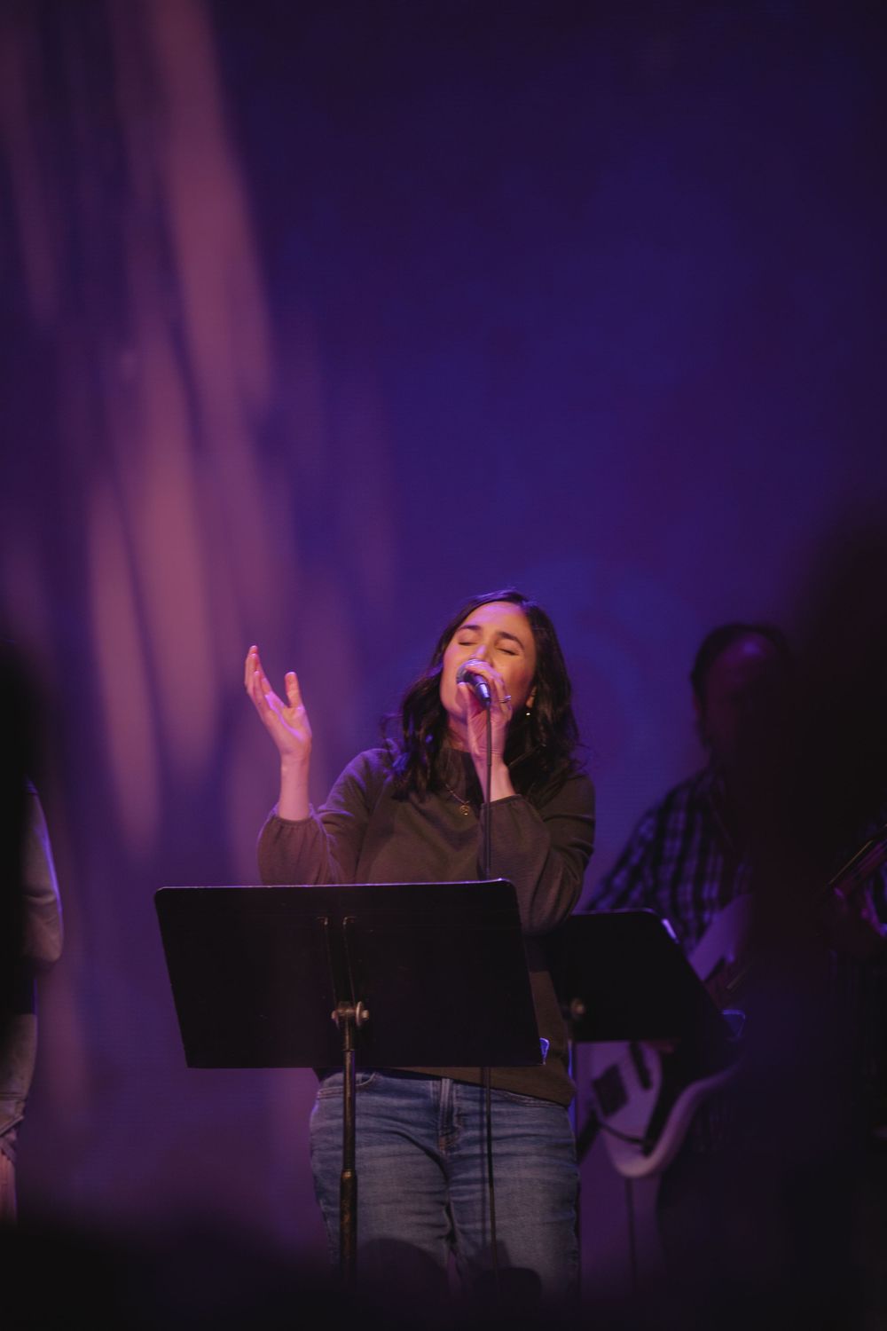 Female worship singer performing passionately on stage with purple lighting and a music stand.