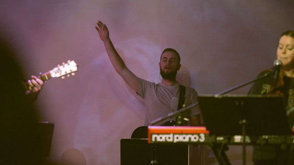 Male worship leader raising one hand while playing keyboard on a foggy lit stage.