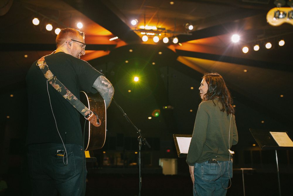 Tattooed guitarist with acoustic guitar talking to woman near microphone stand on stage.