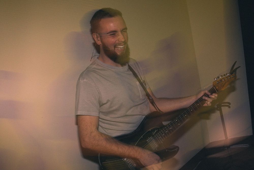 Smiling bearded man playing an electric guitar in warm ambient lighting.