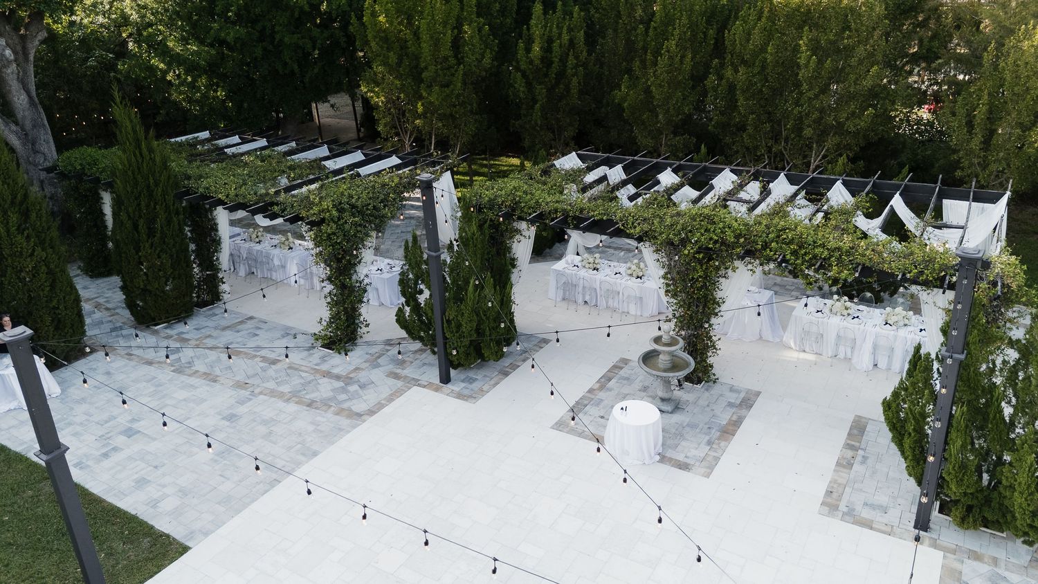 Aerial view of an elegant outdoor wedding venue with white stone flooring, cypress trees, and decorative string lights.