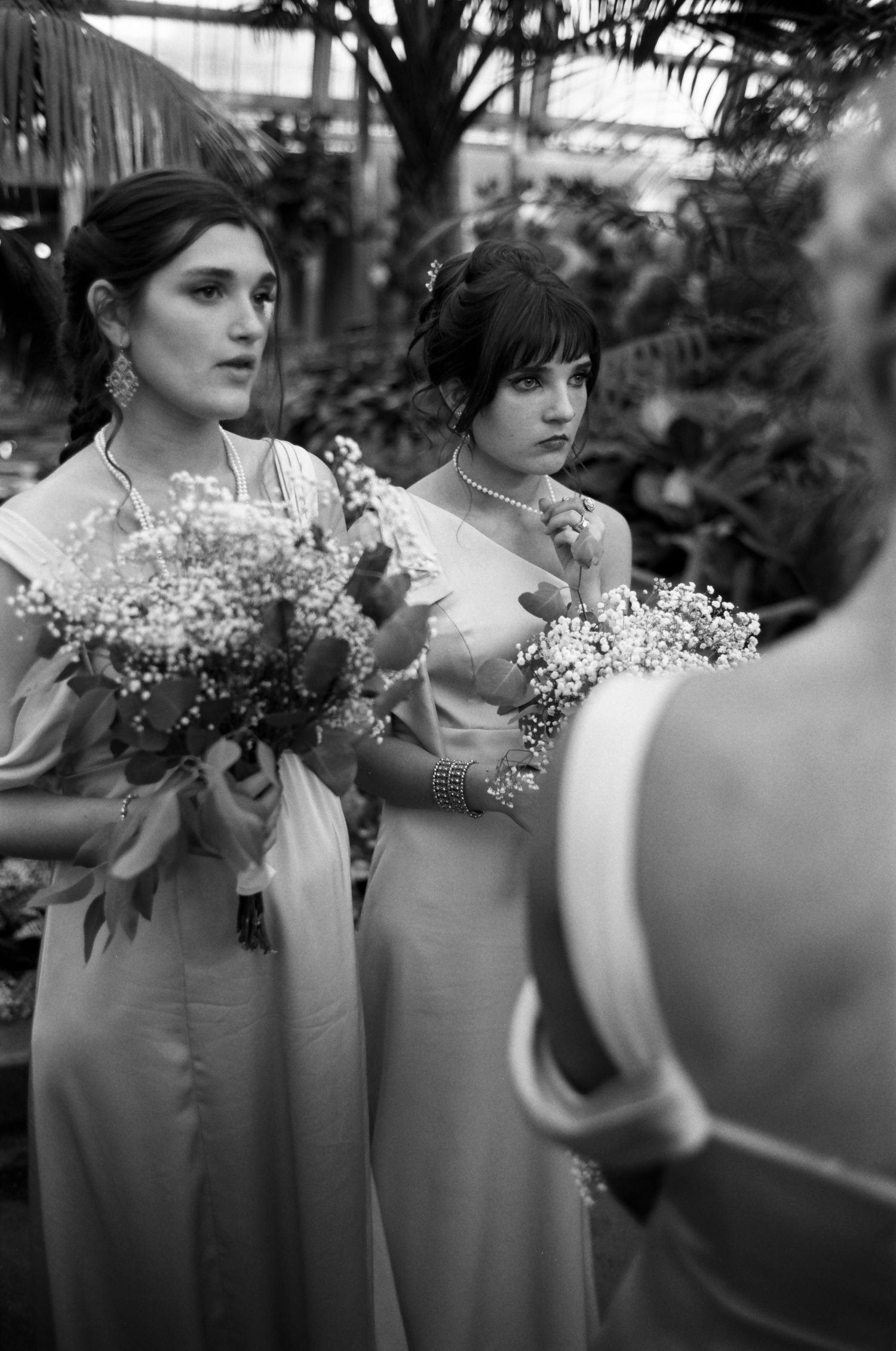 Two bridesmaids in elegant gowns holding floral bouquets at a Midwest greenhouse wedding, black and white analog photo.