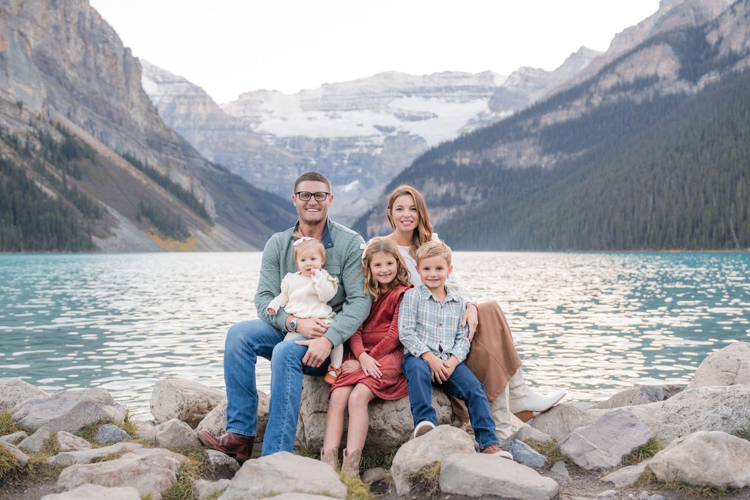 Family of five posing on rocks at turquoise Lake Louise with snow-capped Canadian Rockies mountains in background.