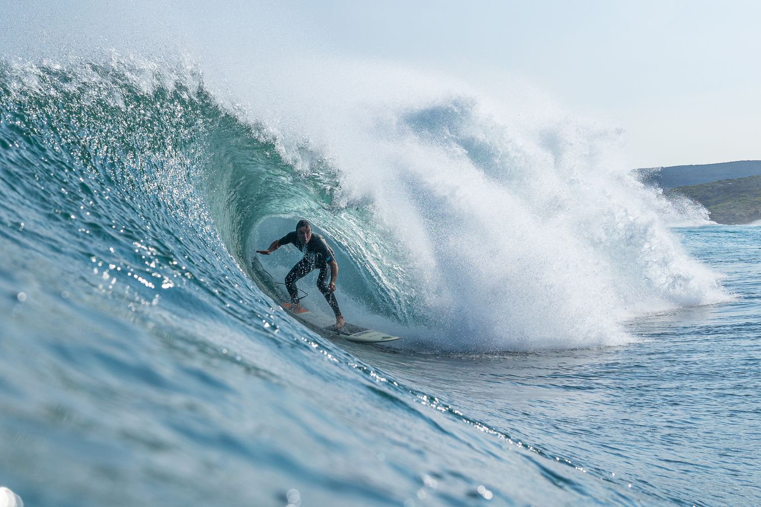 Surfer riding inside a massive barrel wave in the ocean, wearing a wetsuit on a sunny day.