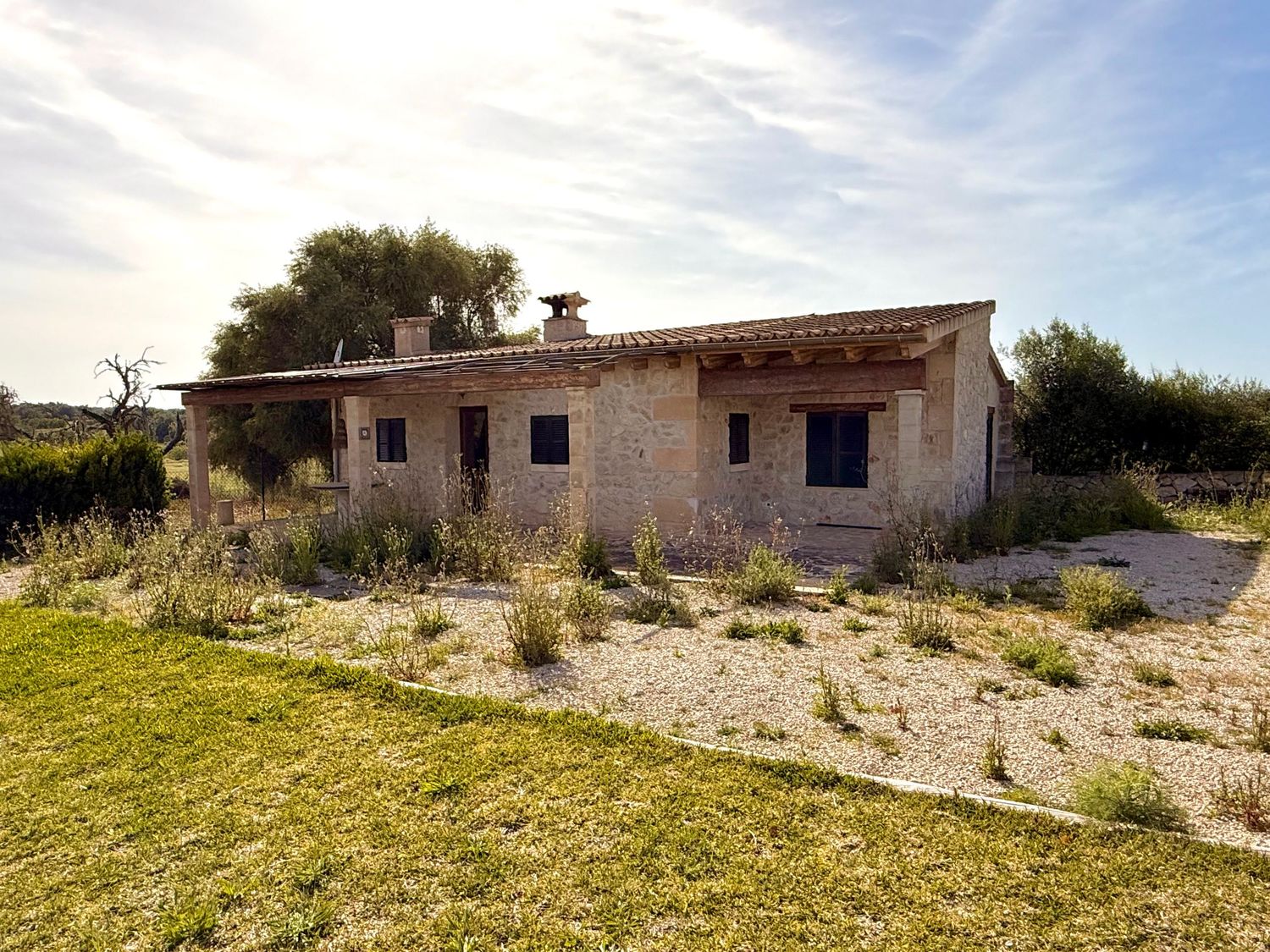 Rustic stone farmhouse with overgrown vegetation, set in a rural landscape under a partly cloudy sky.
