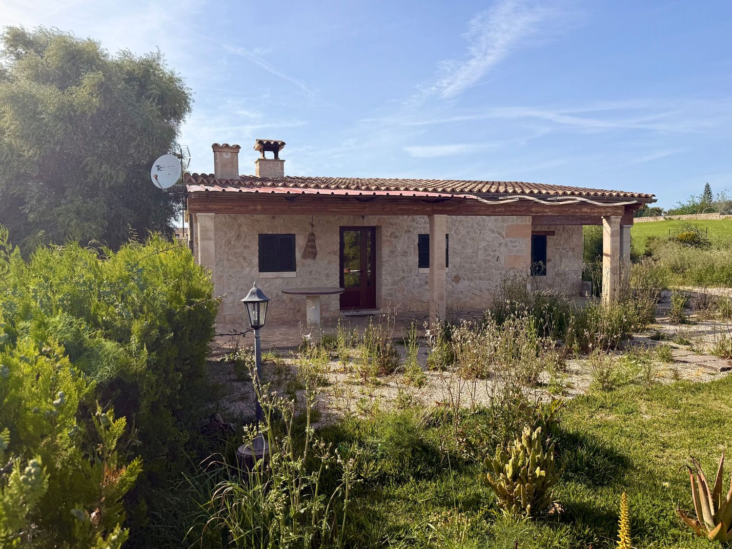 Stone countryside villa with red tile roof, surrounded by lush greenery and natural landscape under a clear blue sky.