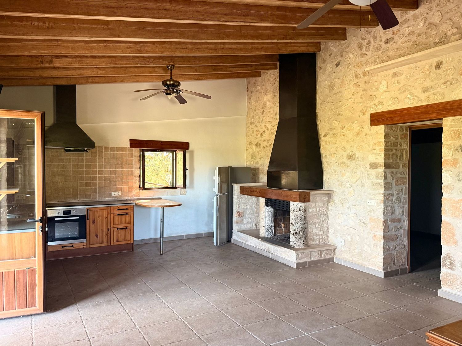 Rustic open-plan kitchen with stone fireplace, wooden ceiling beams, tiled floor, and built-in oven in a Mediterranean-style home.