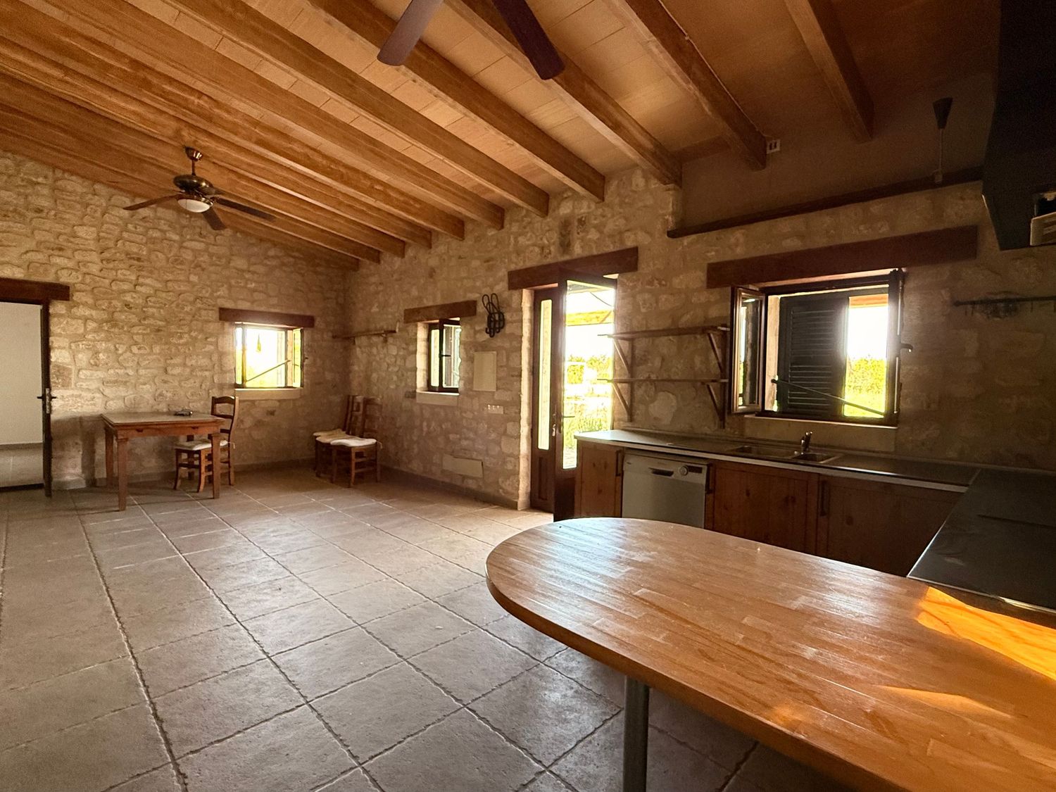 Rustic stone farmhouse interior with wooden beam ceiling, tile floors, kitchen area, and dining space bathed in natural light.