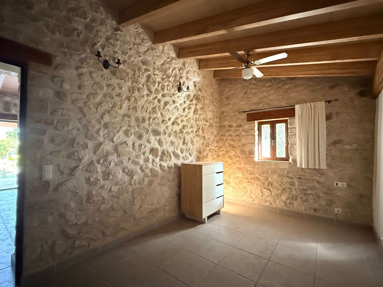 A rustic stone-walled room with wooden beam ceiling, tile floor, dresser, and a ceiling fan.