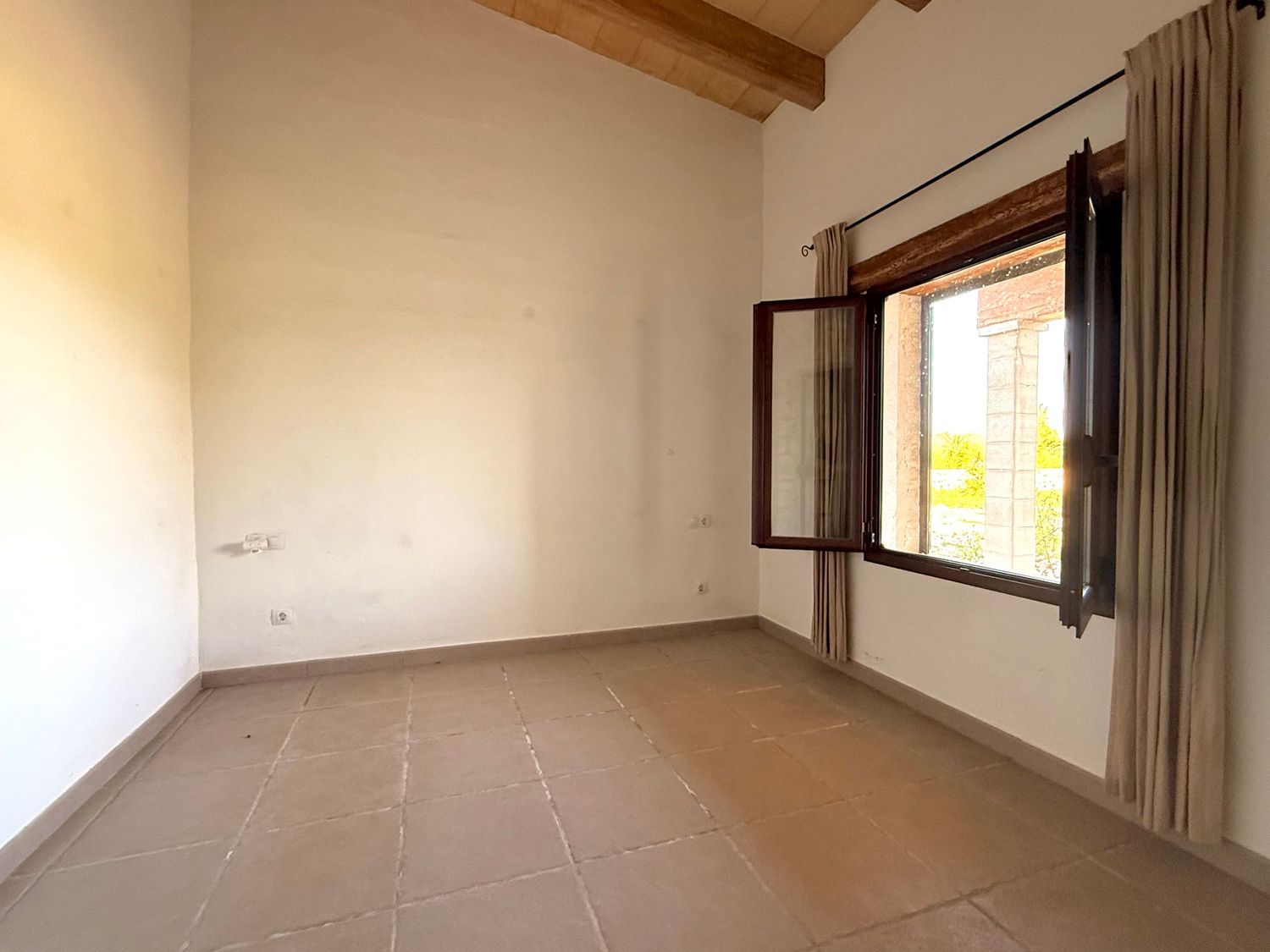 Empty sunlit room with terracotta tile floor, wooden beam ceiling, and open window with curtains.