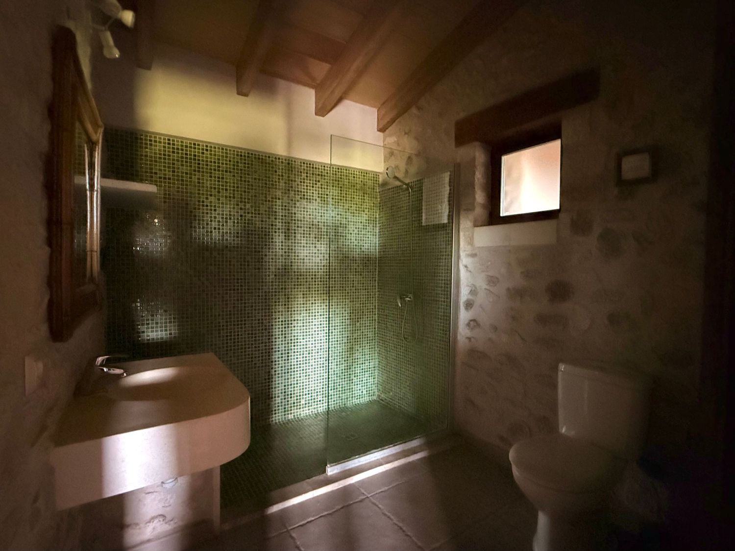 Rustic bathroom with green mosaic tile shower, stone walls, and wooden ceiling beams.