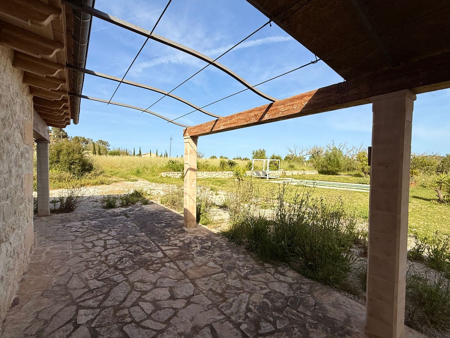 Rustic stone farmhouse patio with pergola overlooking green countryside landscape on sunny day.