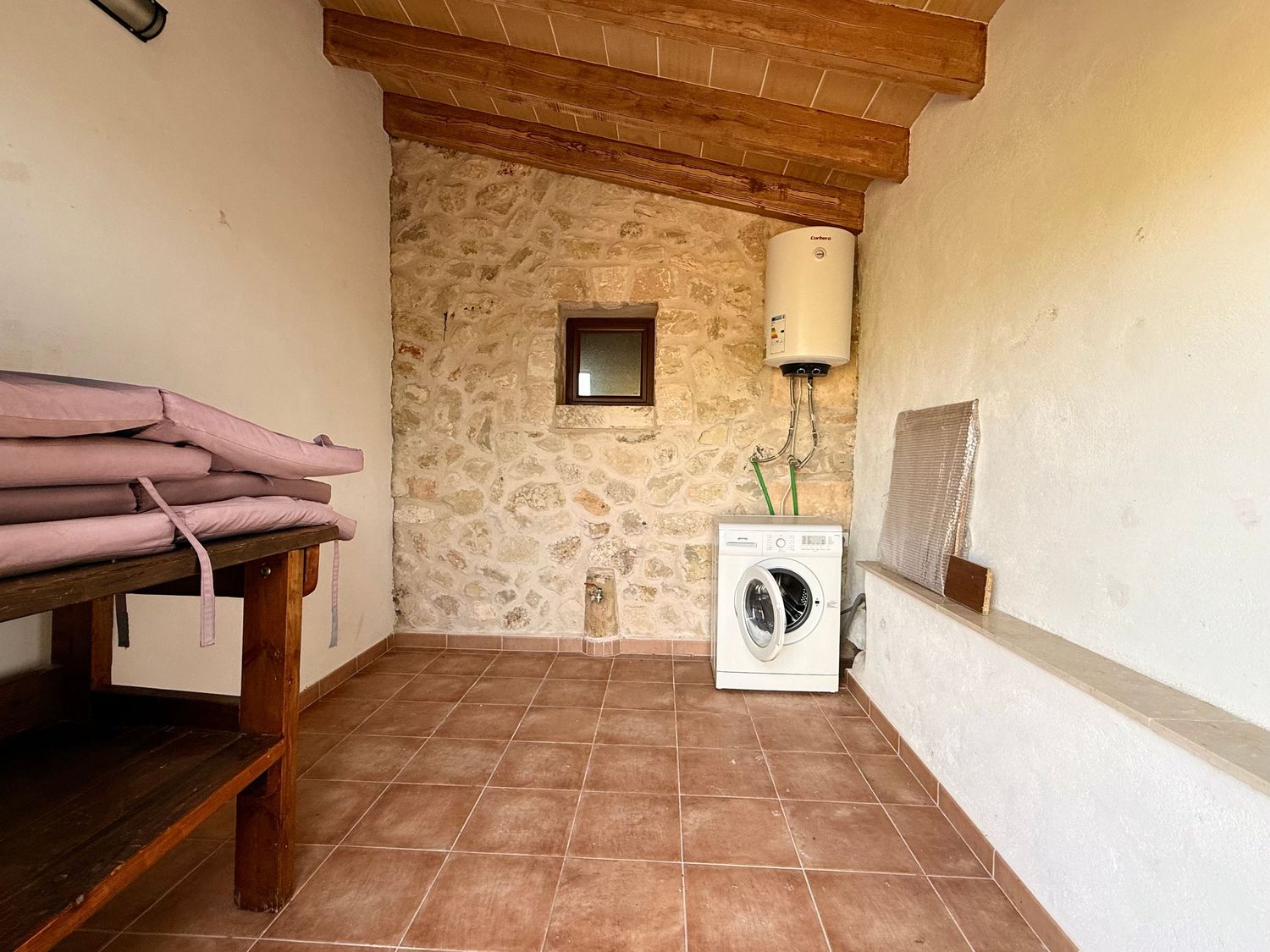 A utility room with stone walls, terracotta tile floor, wooden beams, a washing machine, and water heater.