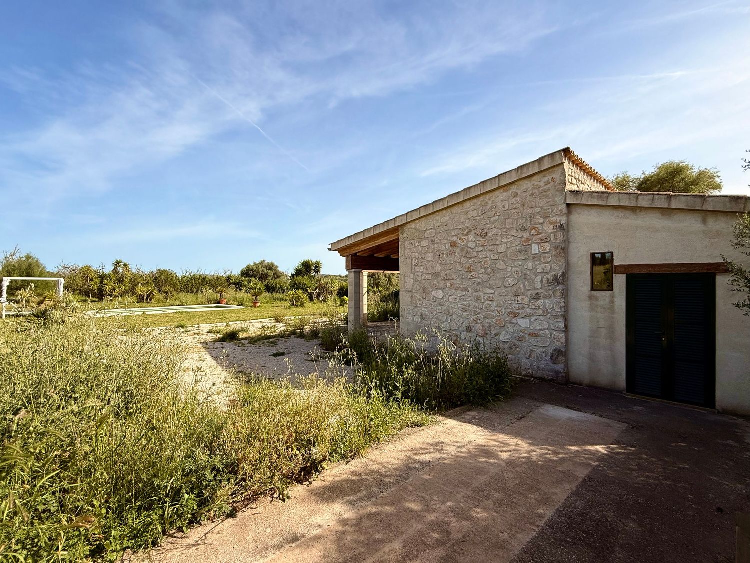 Rustic stone outbuilding with garage door beside a pond under blue sky in a rural countryside setting.
