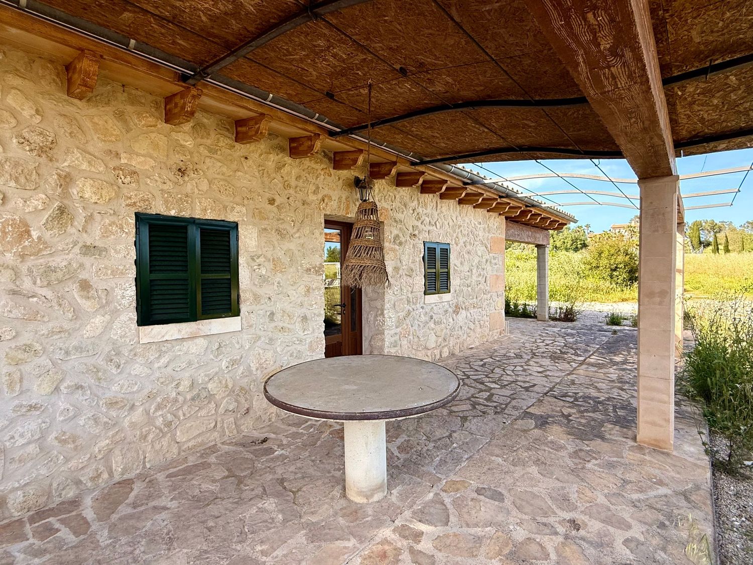 Stone farmhouse patio with round table under wooden pergola, green shuttered window, and scenic countryside views.