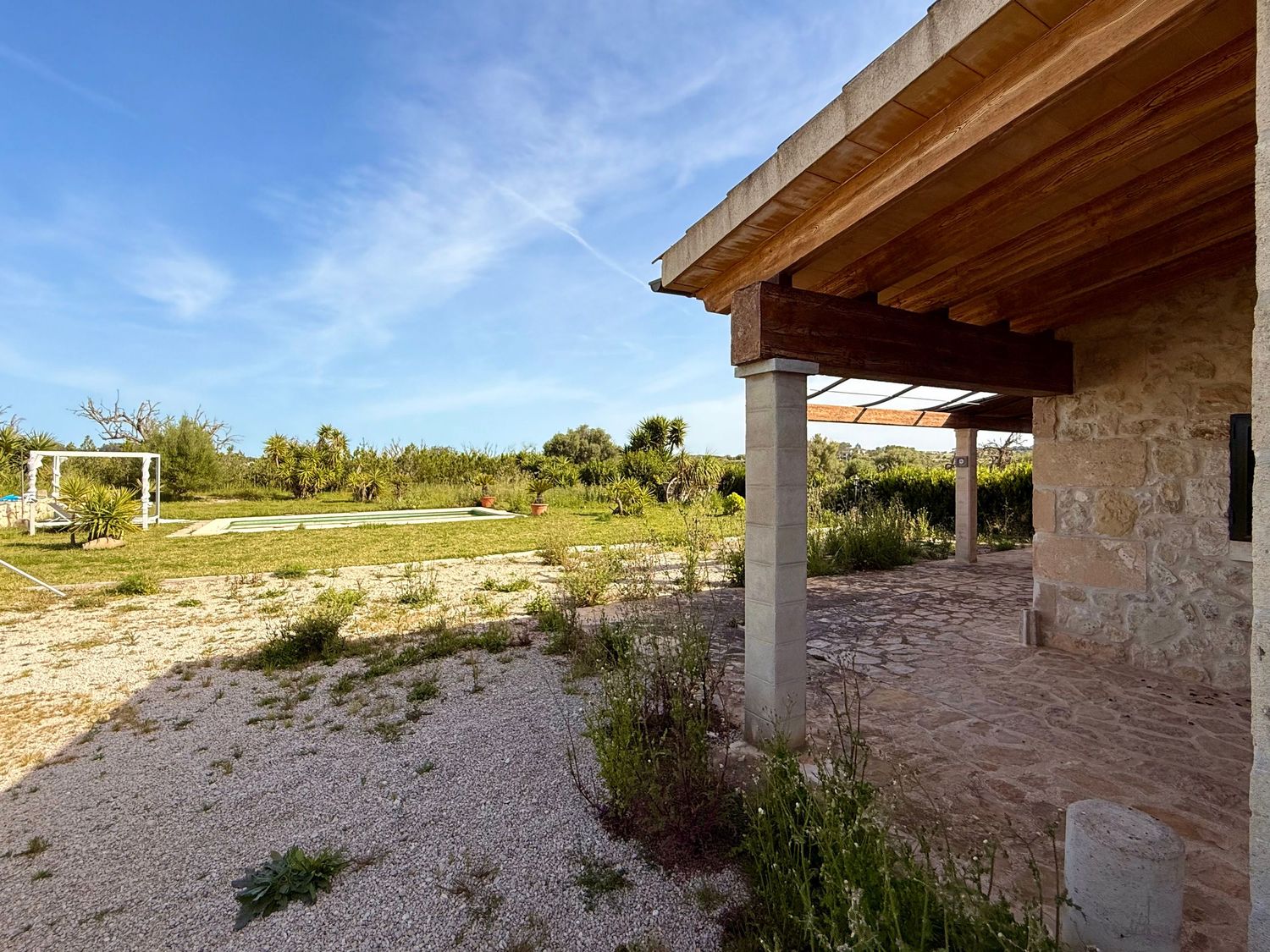 Stone farmhouse with wooden roof terrace overlooking a large rural garden with olive trees under a clear blue sky.