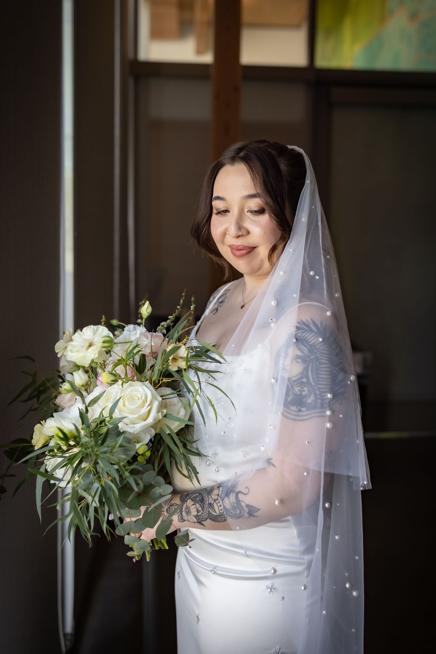 Tattooed bride in white gown and pearl veil smiles while holding elegant white rose bouquet indoors.