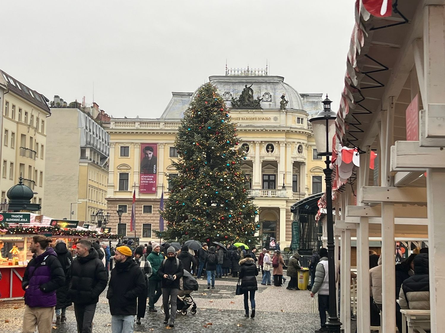 Crowds browse Christmas market stalls surrounding a tall decorated tree in front of a grand European theater building.