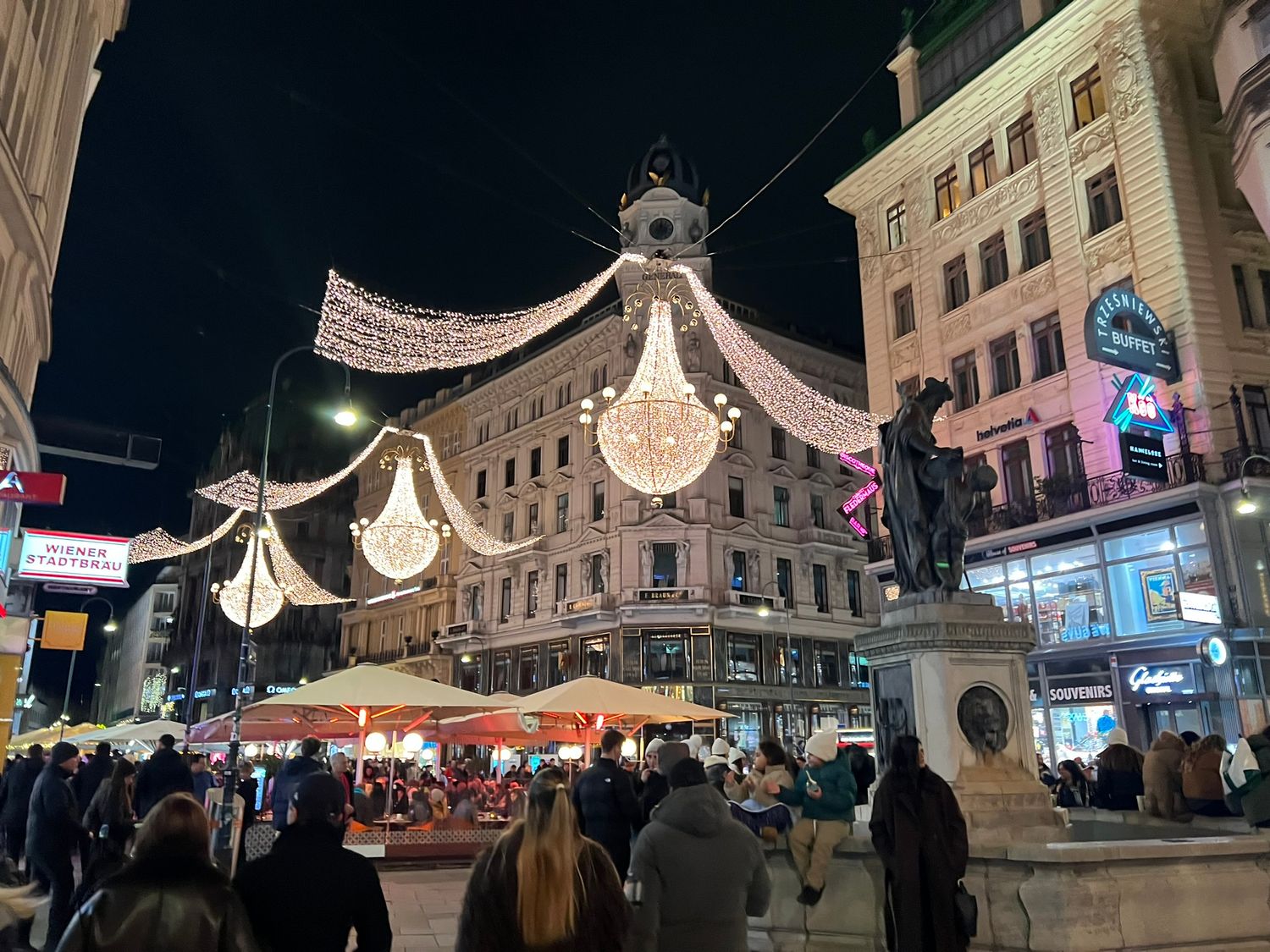 Crowded Vienna street at night decorated with glittering chandelier lights and holiday ornaments above festive outdoor dining.