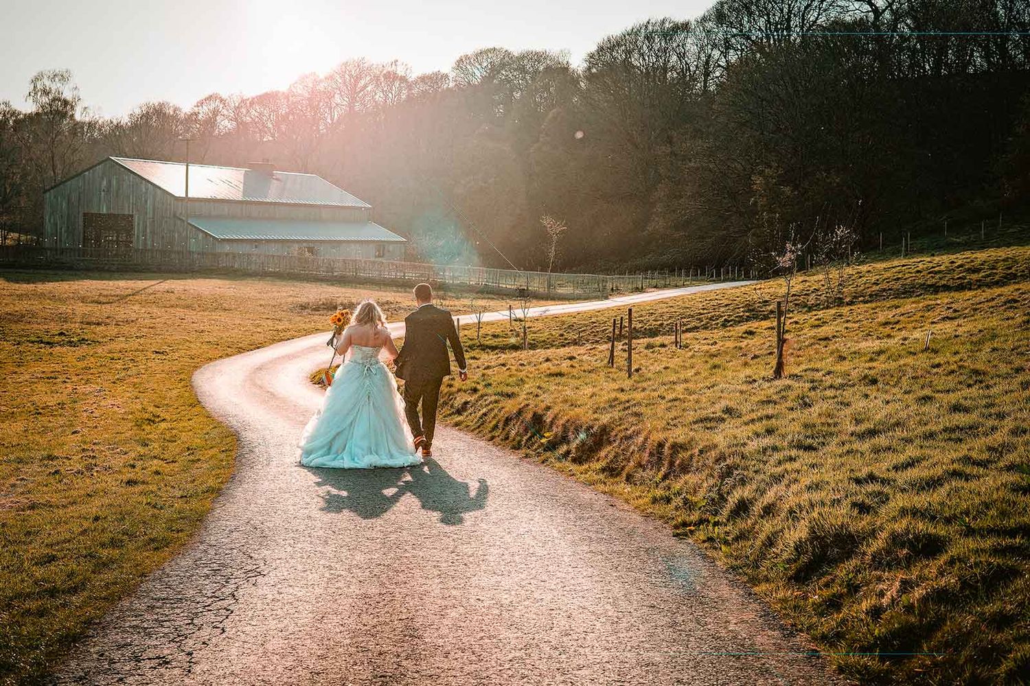 Bride and groom walking hand in hand down a sunlit country path toward a rustic barn on their wedding day.