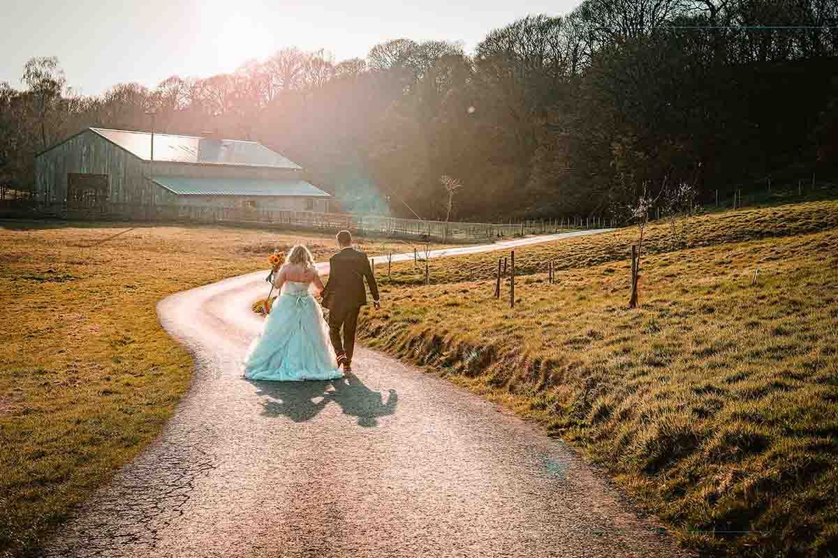 Bride and groom walking hand in hand down a sunlit country path toward a rustic barn on their wedding day.