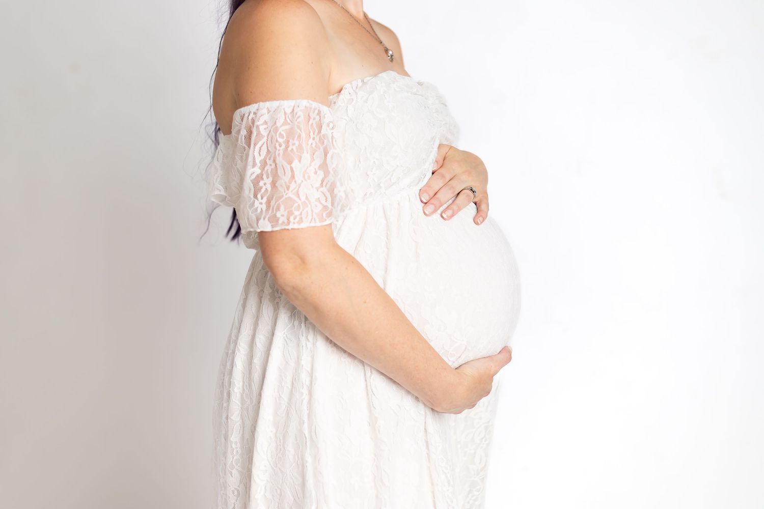 Pregnant woman in white lace off-shoulder dress cradling baby bump against white background.