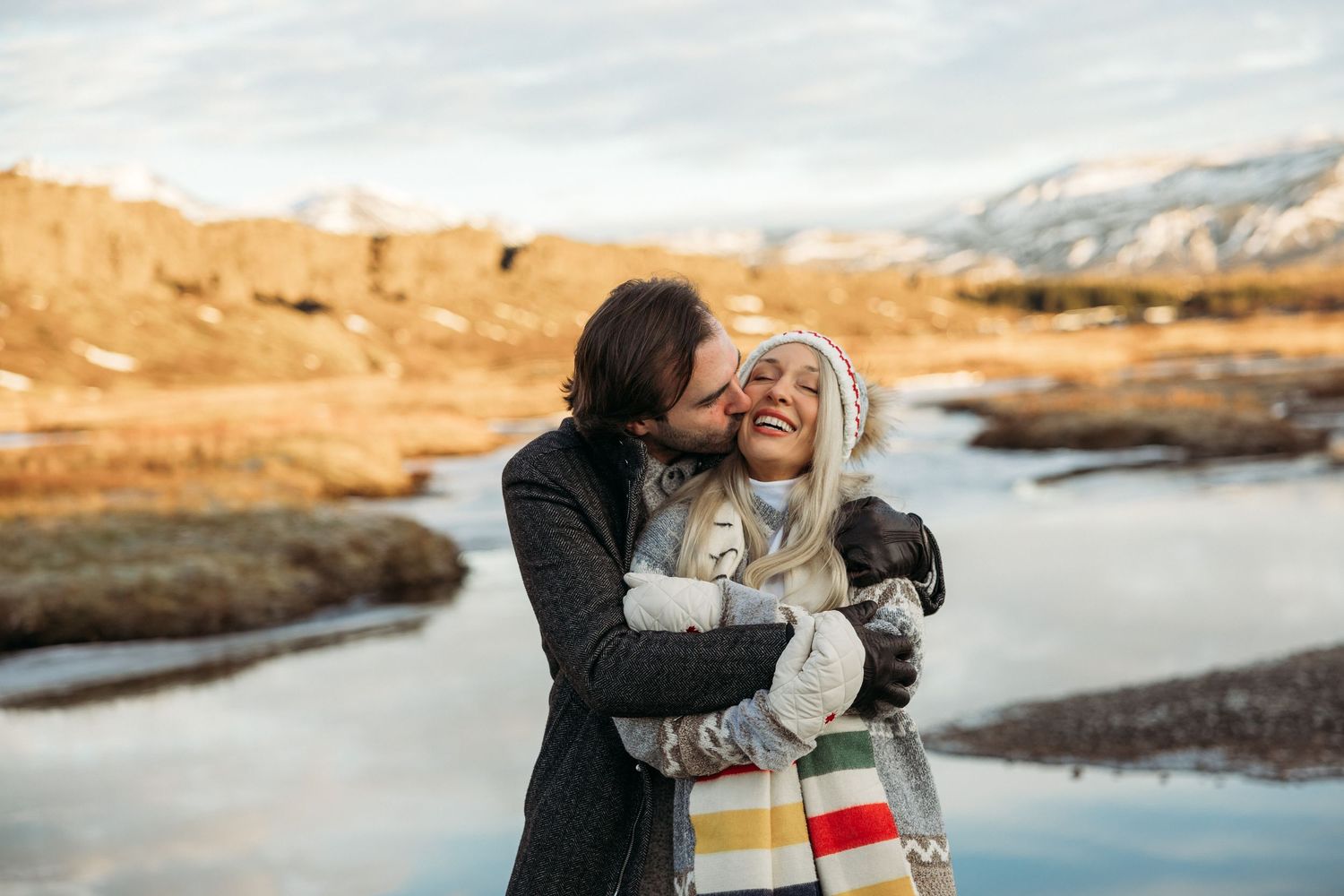 Couple embracing outdoors in winter, woman smiling while man kisses her cheek, holding a dog near a scenic mountain stream.