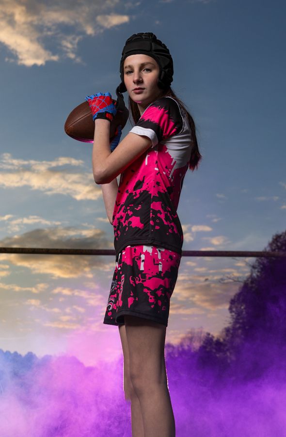 Young female athlete in pink camo uniform holds football, wearing gloves and headgear, with purple smoke in background.