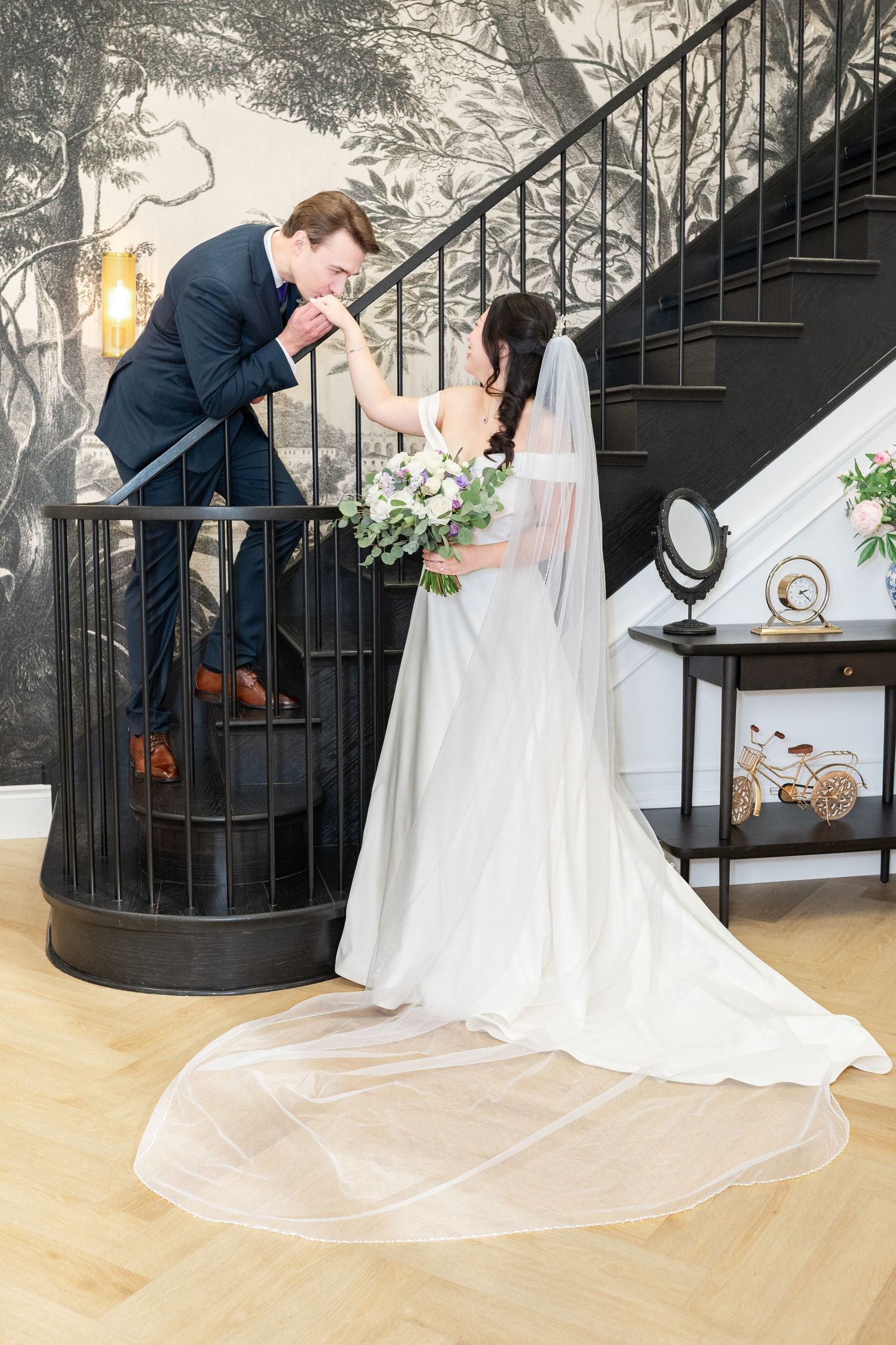 Man in black suit kissing woman's hand in white hijab and gown at formal event.