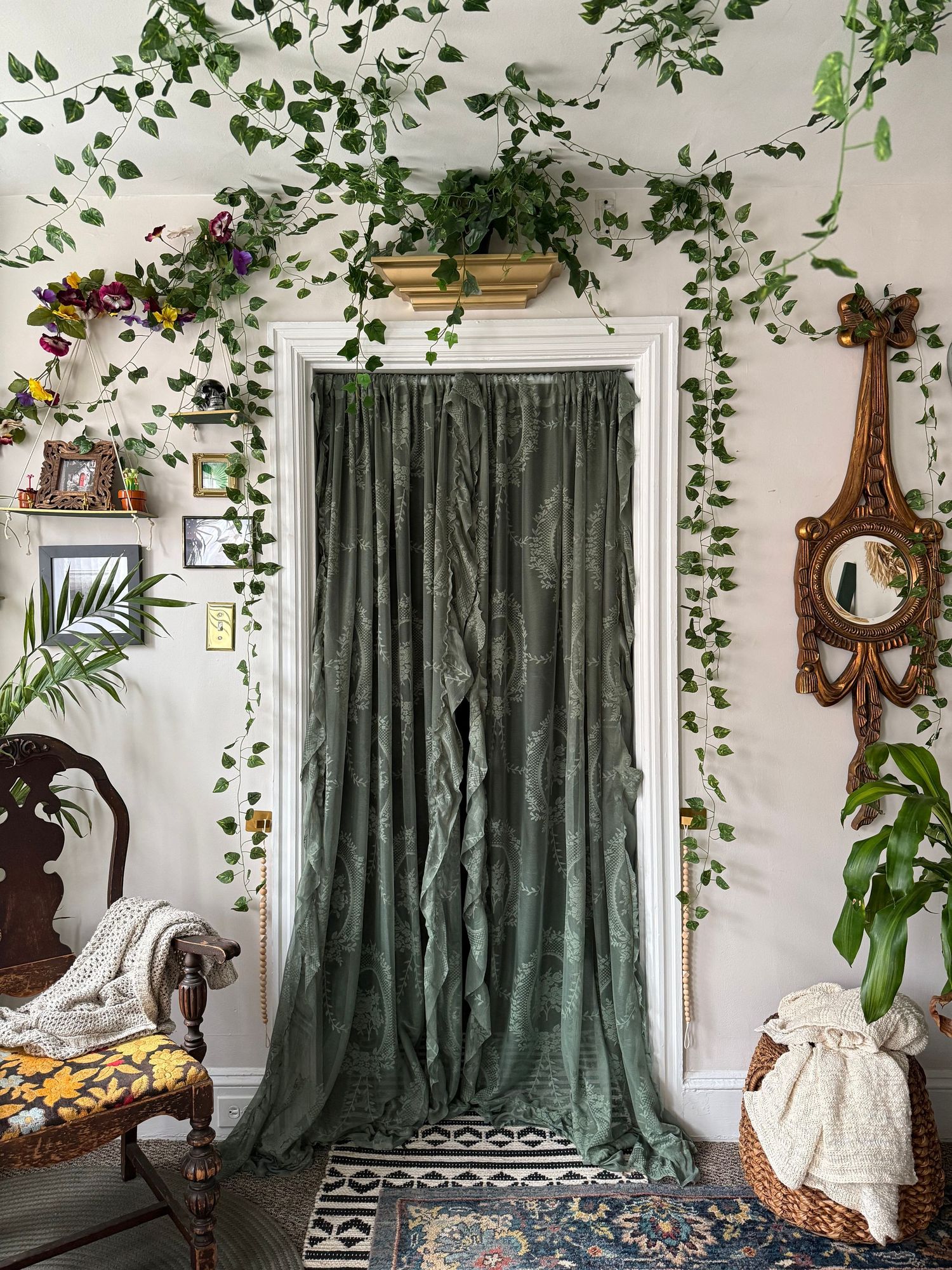 Green velvet curtain doorway adorned with trailing ivy vines, ornate mirror, vintage chair, and lush indoor plants in boho decor.