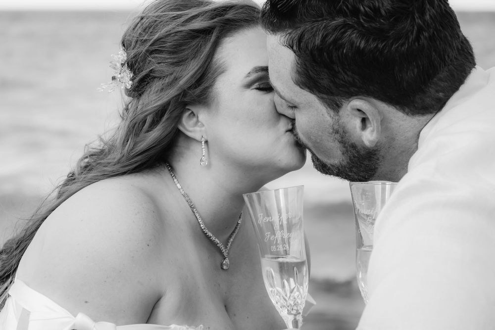 Black and white close-up of bride and groom kissing while holding champagne glasses at beach wedding.