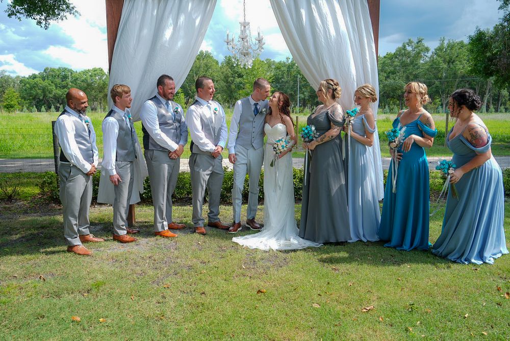 Wedding party in gray suits and blue bridesmaid gowns posing under white arch at outdoor ceremony.