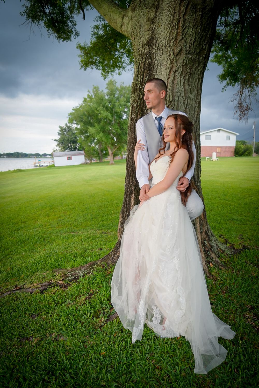 Bride in flowing white gown and groom in gray suit posing together under large tree by lakeside.