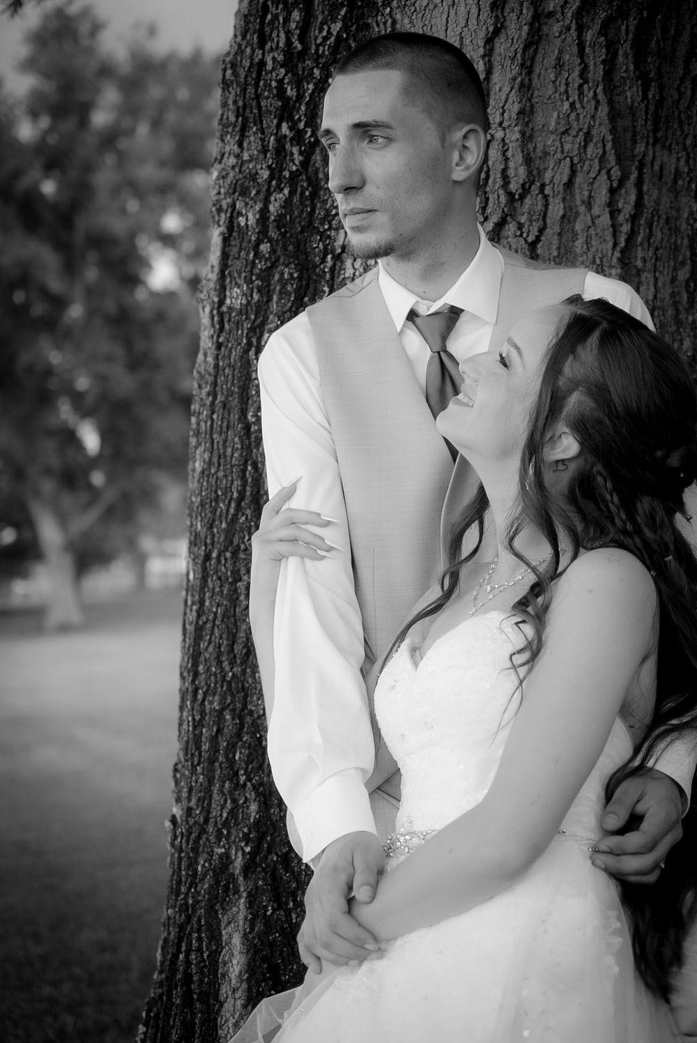 Black and white portrait of bride and groom embracing against large tree trunk at outdoor wedding.