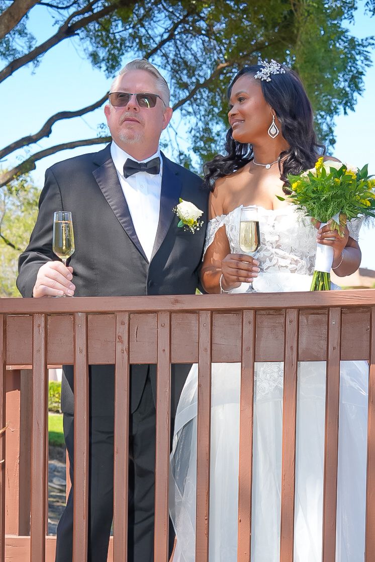 Interracial couple in wedding attire toasting champagne on wooden deck surrounded by trees.